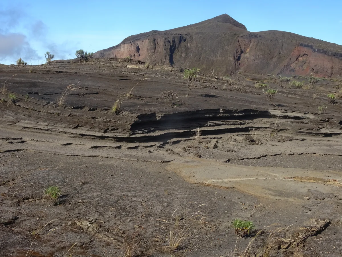 Exposed tephra layers and the summit ridge of Mount Tambora under a blue sky